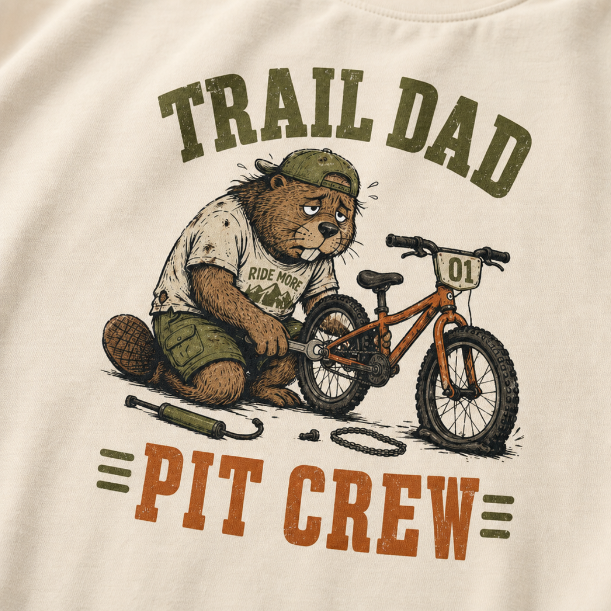 Close-up of an ivory white t-shirt with a funny “Trail Dad Pit Crew” design showing an exhausted beaver repairing a small mountain bike.