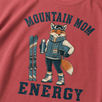 Close-up of a crimson red t-shirt with a retro “Mountain Mom Energy” ski design showing a confident fox holding a thermos and helmet beside a pair of skis.