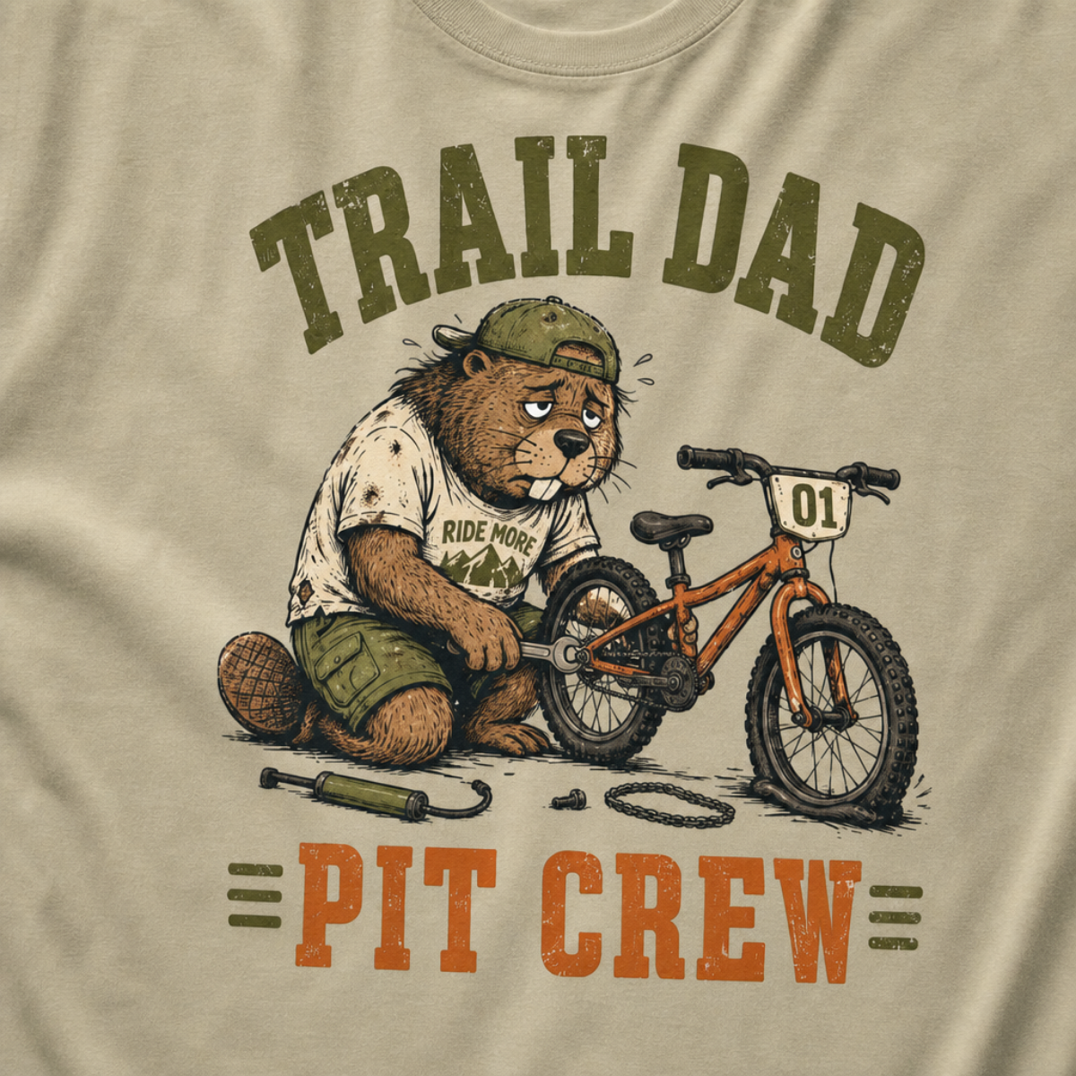 Close-up of a khaki t-shirt with a funny “Trail Dad Pit Crew” design showing an exhausted beaver repairing a small mountain bike.