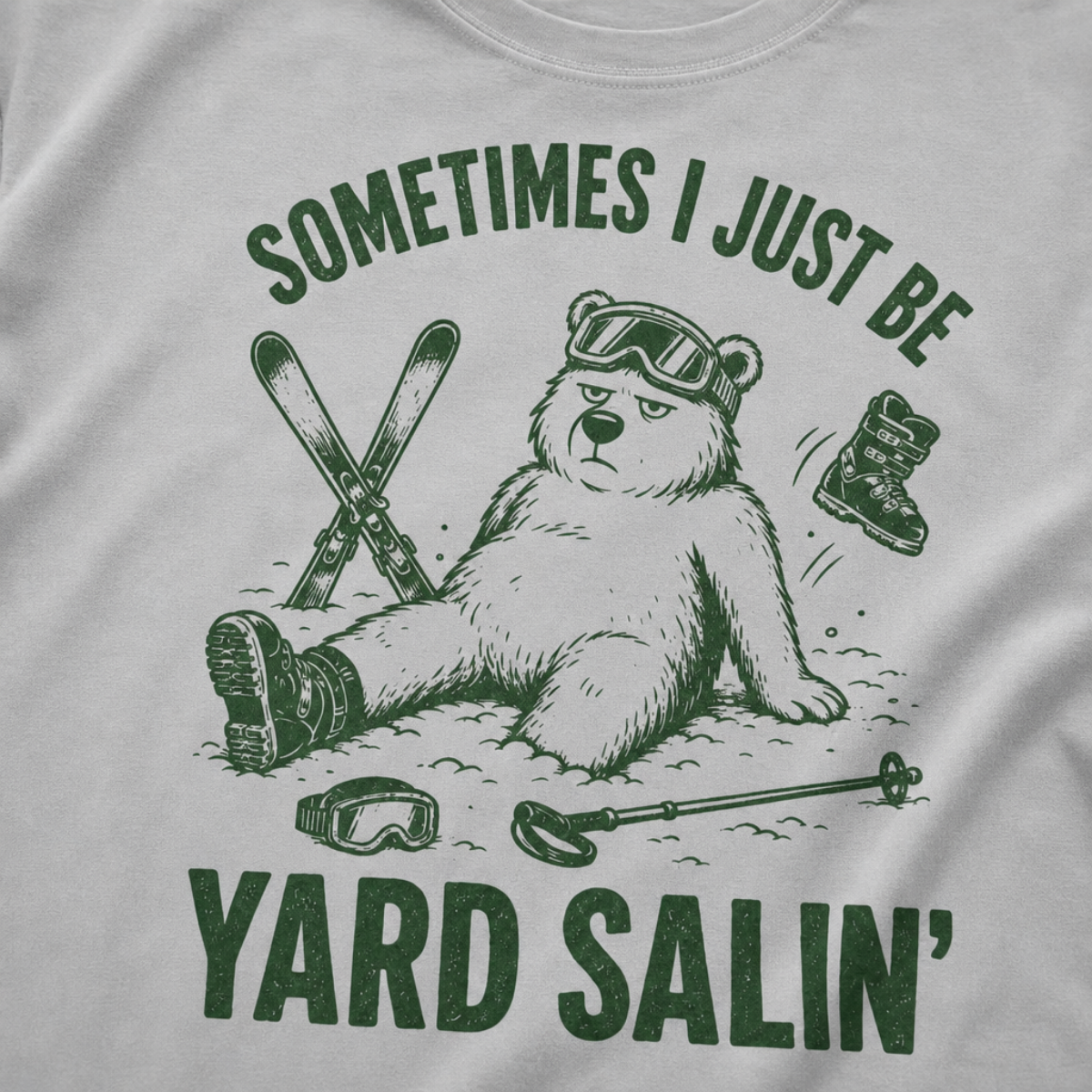 Close-up of a granite gray t-shirt with a green vintage ski graphic showing a worn-out bear after a yard sale crash, surrounded by skis, goggles, poles, and a boot.