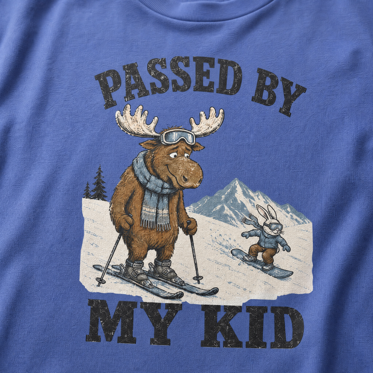 Close-up of a mystic blue t-shirt with a vintage “Passed By My Kid” design showing an embarrassed moose skier being passed by a tiny rabbit snowboarder.