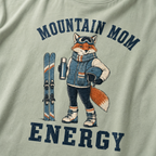 Close-up of a bay green t-shirt with a retro “Mountain Mom Energy” ski design showing a confident fox holding a thermos and helmet beside a pair of skis.