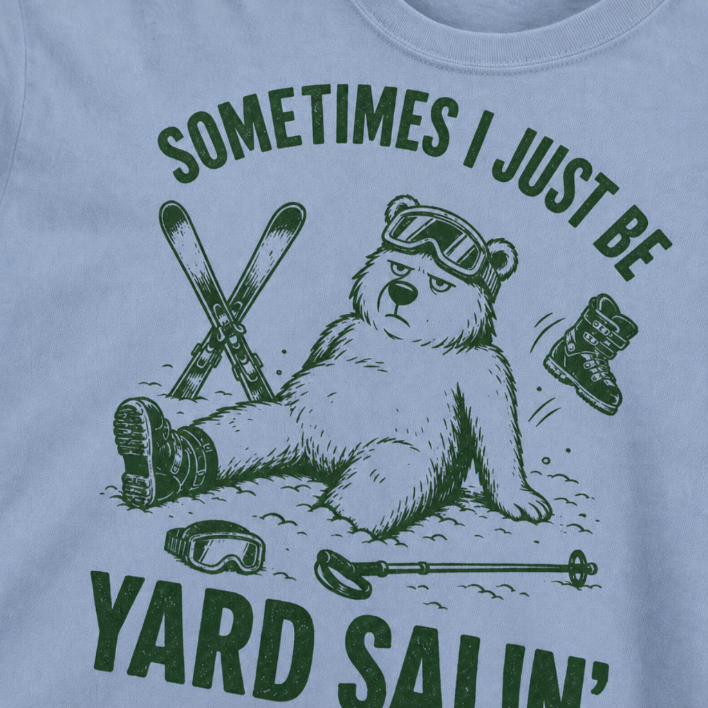 Close-up of a washed denim t-shirt with a green vintage ski graphic showing a worn-out bear after a yard sale crash, surrounded by skis, goggles, poles, and a boot.