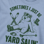 Close-up of a washed denim t-shirt with a green vintage ski graphic showing a worn-out bear after a yard sale crash, surrounded by skis, goggles, poles, and a boot.