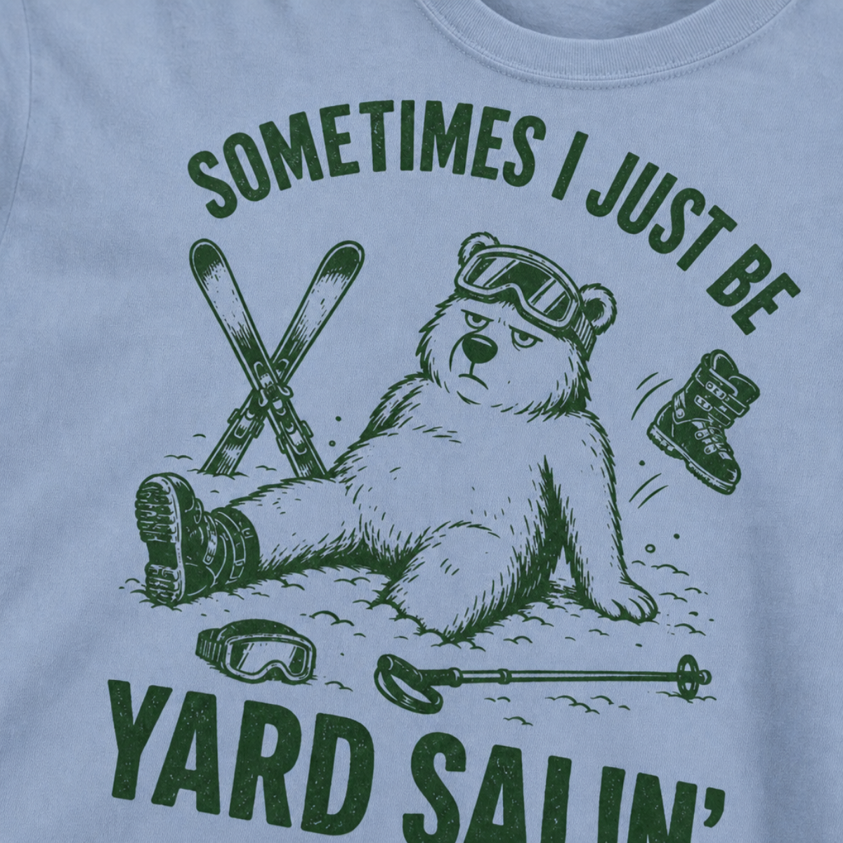 Close-up of a washed denim t-shirt with a green vintage ski graphic showing a worn-out bear after a yard sale crash, surrounded by skis, goggles, poles, and a boot.