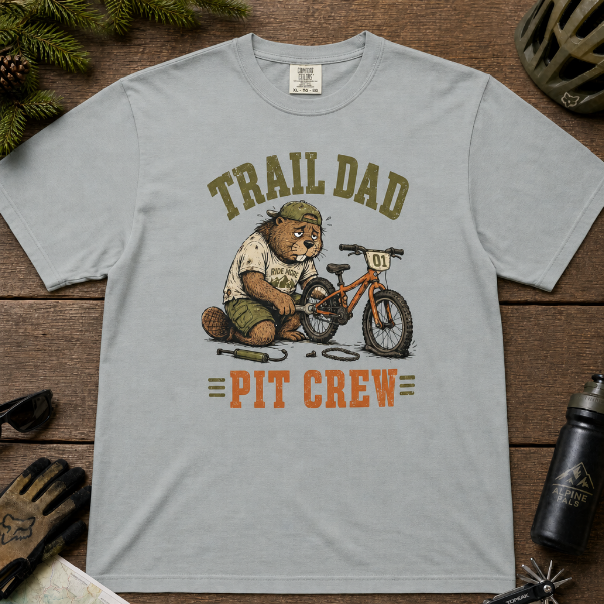Flat lay of a granite gray t-shirt featuring a “Trail Dad Pit Crew” mountain biking design with a tired beaver fixing a kid’s bike, styled with trail accessories.