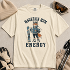 Flat lay of an ivory white t-shirt featuring a “Mountain Mom Energy” fox ski graphic, styled with winter accessories like goggles, gloves, and poles.