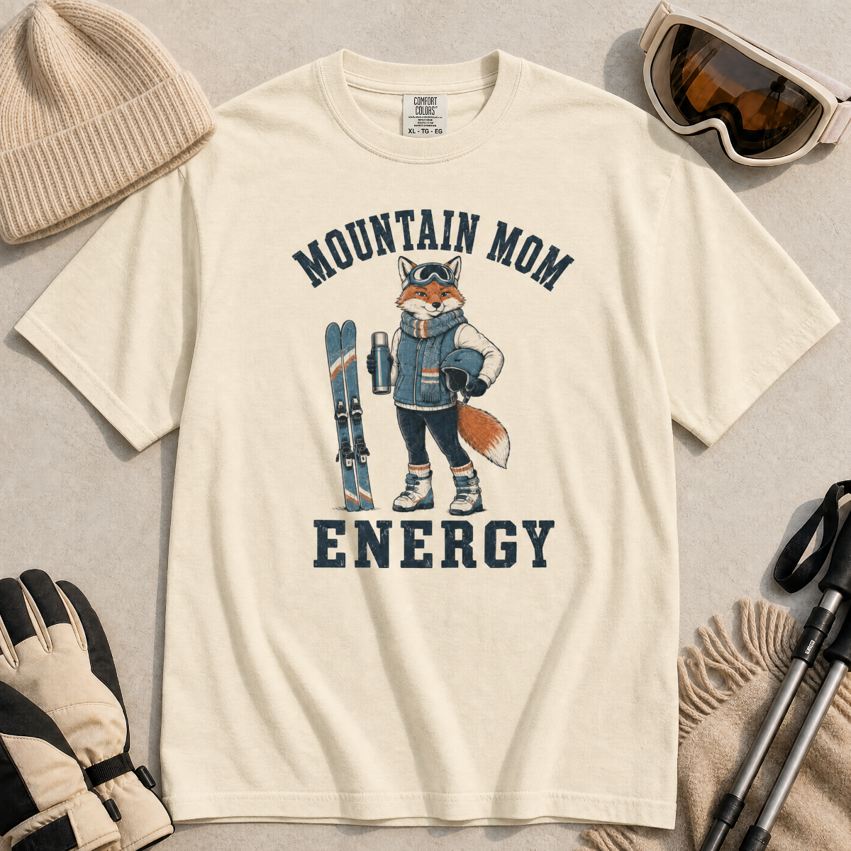 Flat lay of an ivory white t-shirt featuring a “Mountain Mom Energy” fox ski graphic, styled with winter accessories like goggles, gloves, and poles.