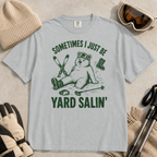Flat lay of a granite gray t-shirt featuring a funny “Sometimes I Just Be Yard Salin’” ski design with a tired bear sitting in the snow among scattered gear.