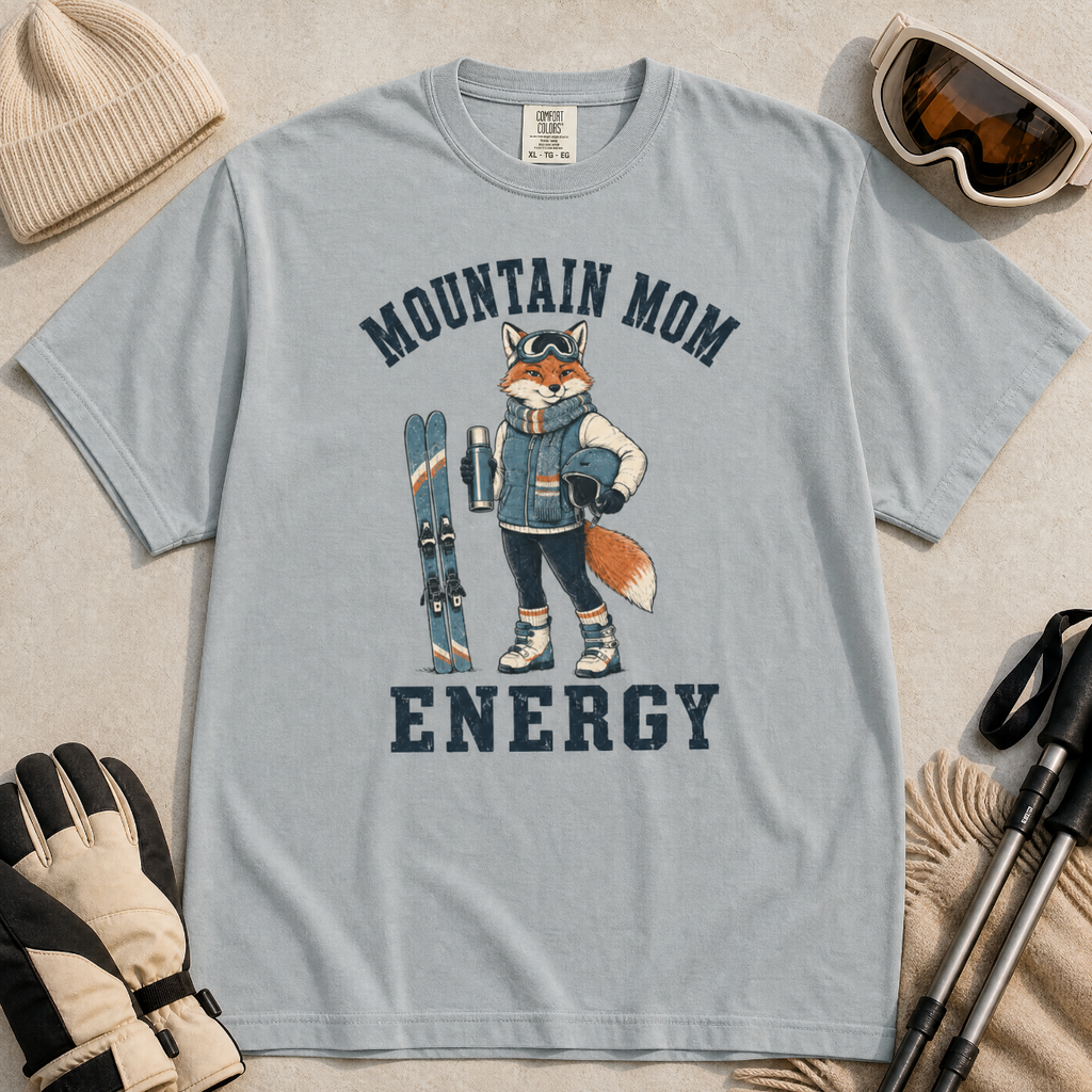 Flat lay of a granite gray t-shirt featuring a “Mountain Mom Energy” fox ski graphic, styled with winter accessories like goggles, gloves, and poles.
