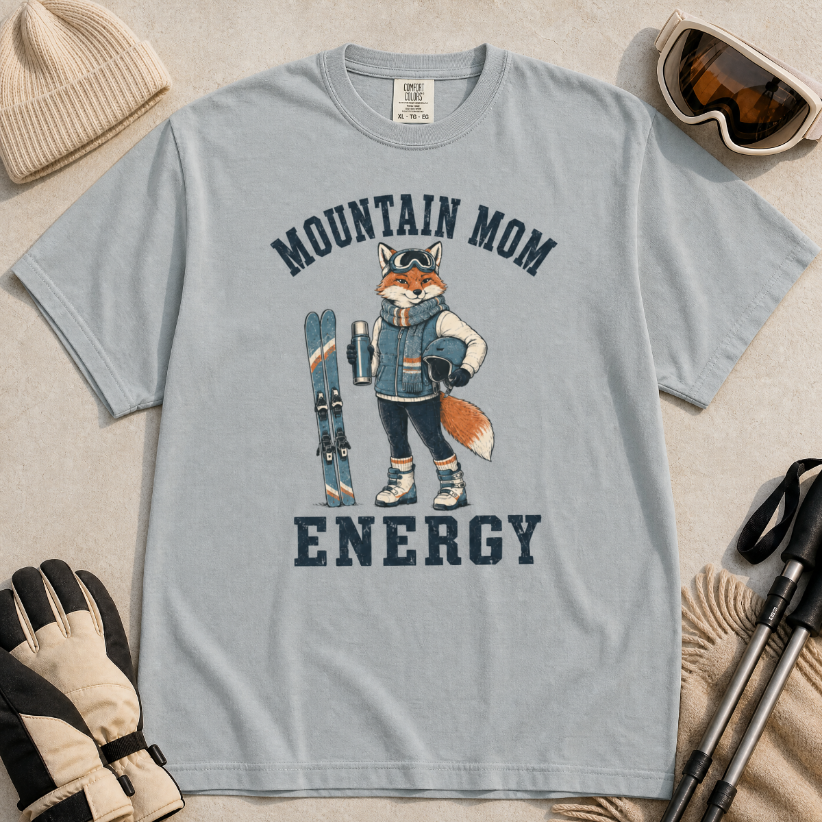 Flat lay of a granite gray t-shirt featuring a “Mountain Mom Energy” fox ski graphic, styled with winter accessories like goggles, gloves, and poles.