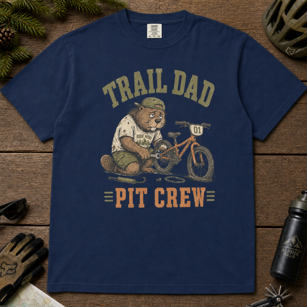 Flat lay of a navy t-shirt featuring a “Trail Dad Pit Crew” mountain biking design with a tired beaver fixing a kid’s bike, styled with trail accessories.