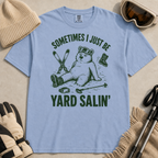 Flat lay of a washed denim blue t-shirt featuring a funny “Sometimes I Just Be Yard Salin’” ski design with a tired bear sitting in the snow among scattered gear.