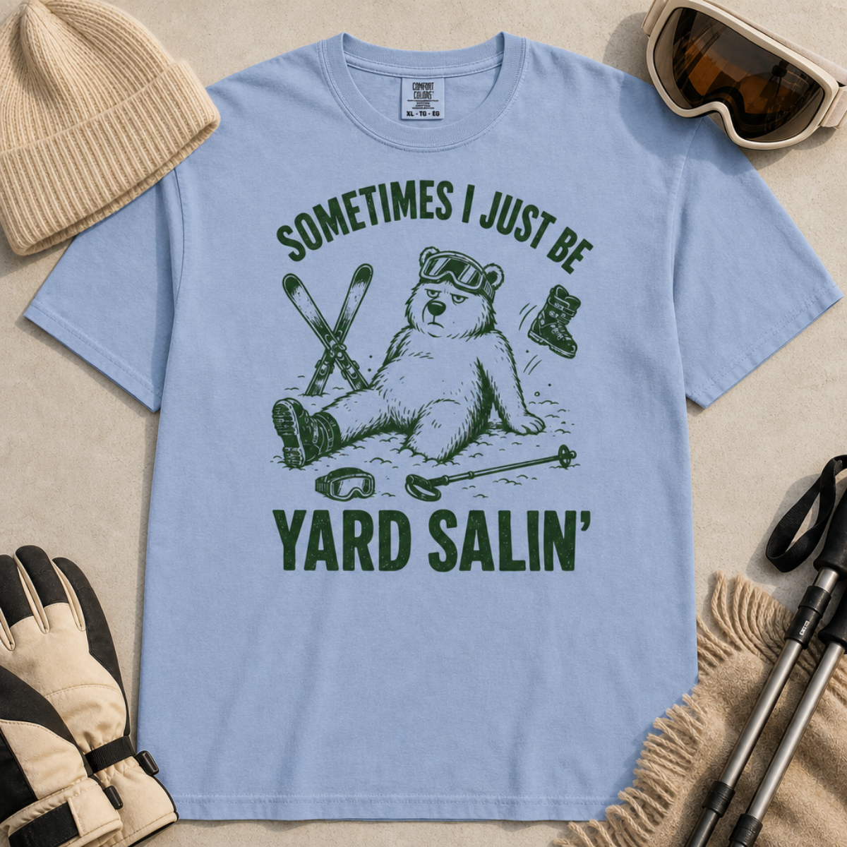 Flat lay of a washed denim blue t-shirt featuring a funny “Sometimes I Just Be Yard Salin’” ski design with a tired bear sitting in the snow among scattered gear.