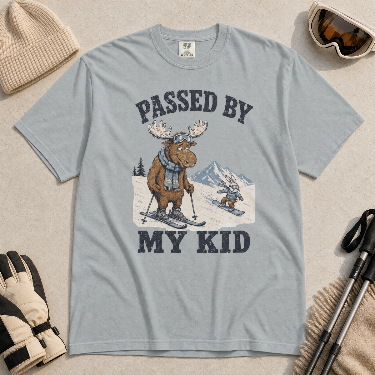 Flat lay of a granite gray t-shirt featuring a funny “Passed By My Kid” ski design with a humbled moose watching a small rabbit snowboarder pass by.