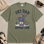 Flat lay of a moss green t-shirt featuring a tired bear carrying skis and ski gear, with “Ski Dad Support Crew” text.