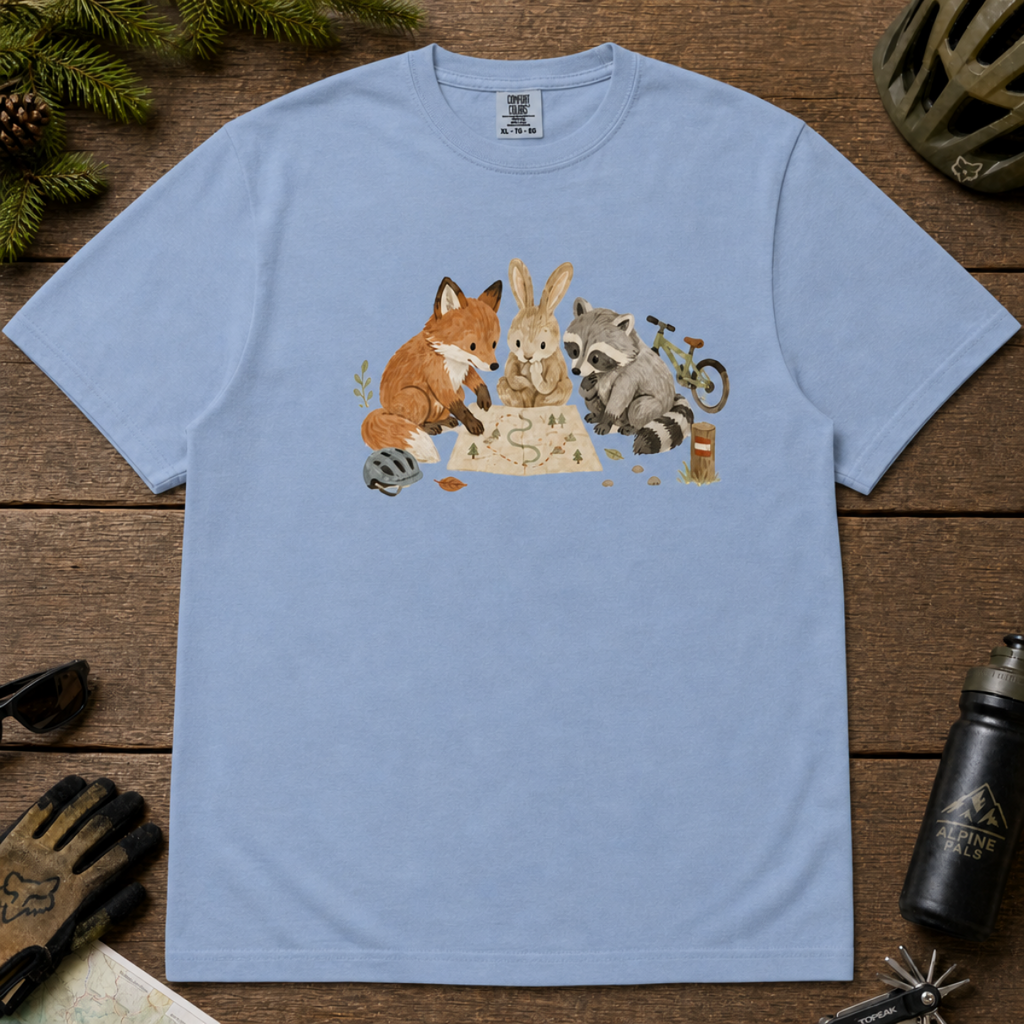 Flat lay of a washed denim blue t-shirt featuring a cute woodland animal trail-planning graphic with a fox, rabbit, raccoon, map, helmet, bike, and trail marker.