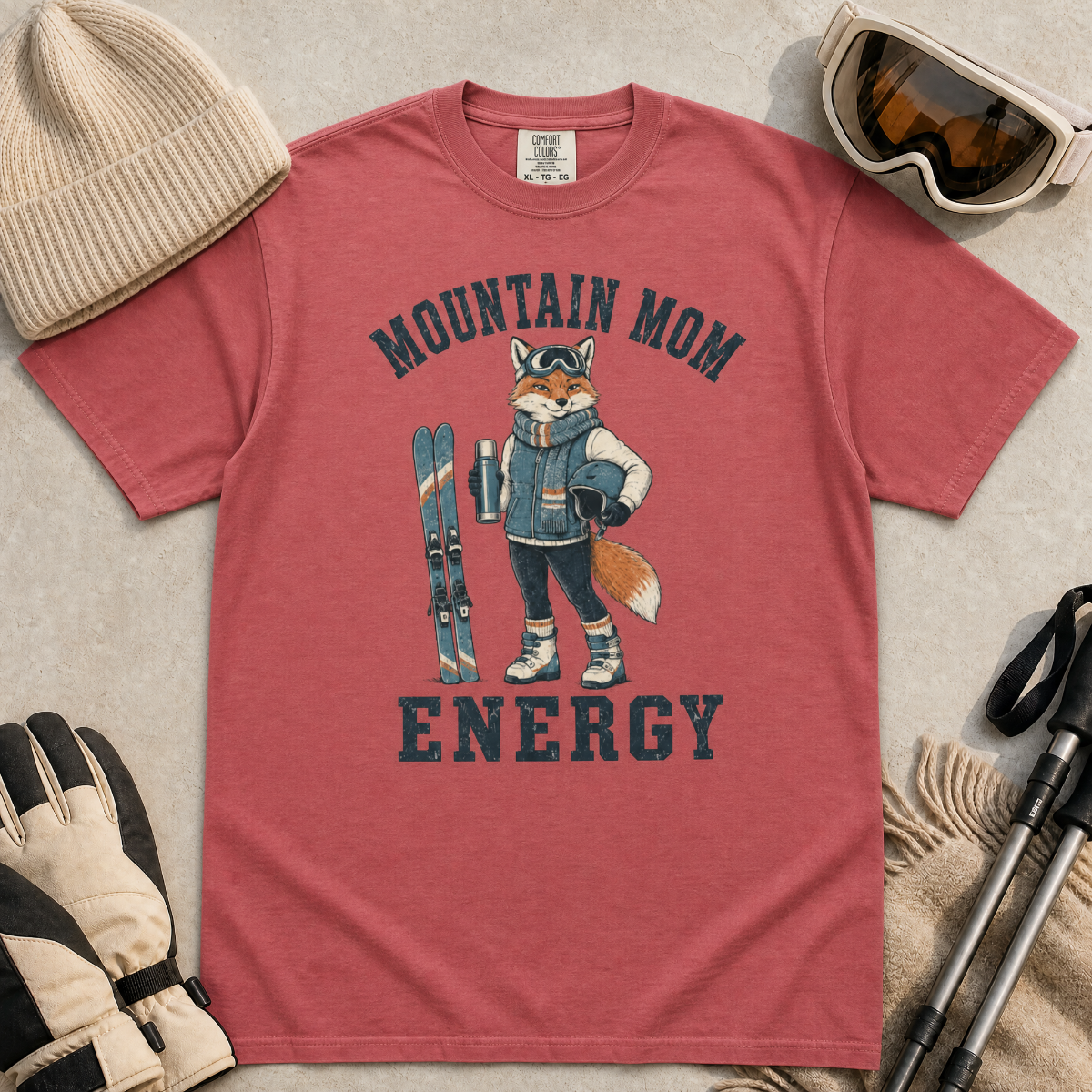 Flat lay of a crimson red t-shirt featuring a “Mountain Mom Energy” fox ski graphic, styled with winter accessories like goggles, gloves, and poles.