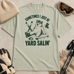 Flat lay of a bay green t-shirt featuring a funny “Sometimes I Just Be Yard Salin’” ski design with a tired bear sitting in the snow among scattered gear.