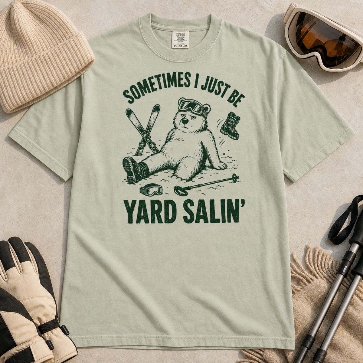 Flat lay of a bay green t-shirt featuring a funny “Sometimes I Just Be Yard Salin’” ski design with a tired bear sitting in the snow among scattered gear.