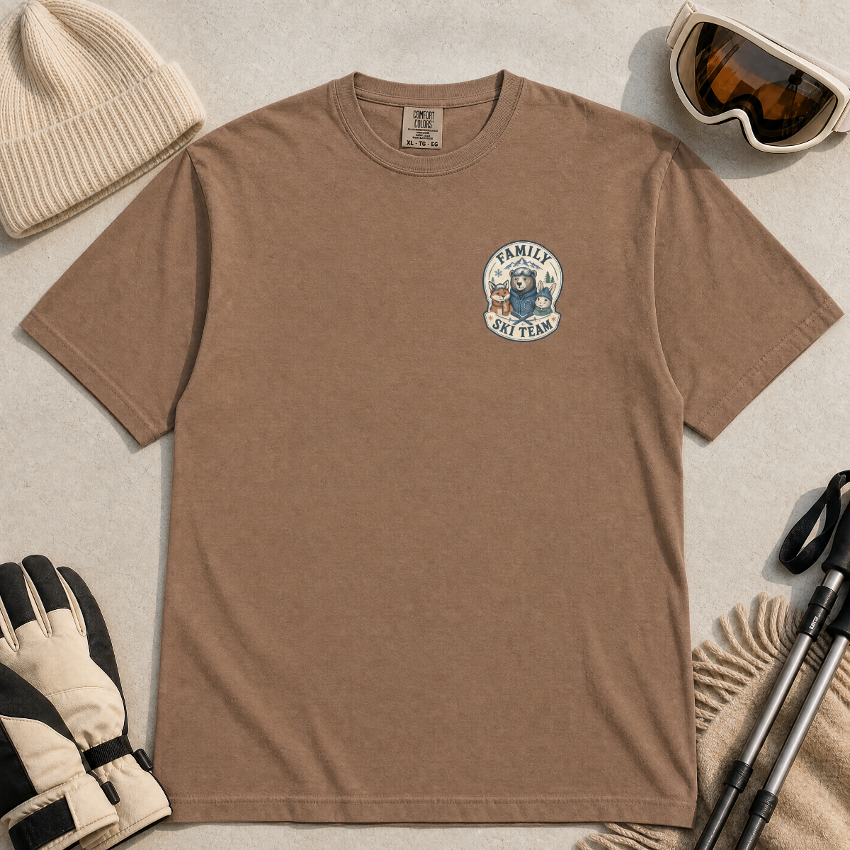 Flat lay of a espresso brown t-shirt featuring a small chest “Family Ski Team” badge graphic with three winter animal characters, styled with ski accessories.