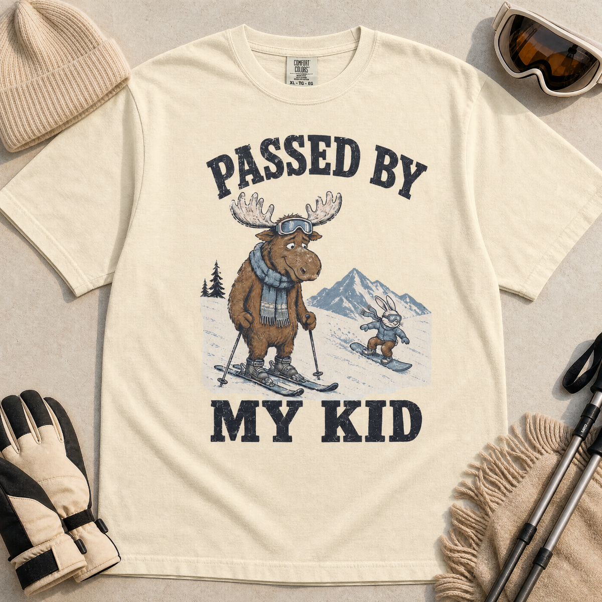 Flat lay of a ivroy white t-shirt featuring a funny “Passed By My Kid” ski design with a humbled moose watching a small rabbit snowboarder pass by.