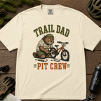 Flat lay of an ivory white t-shirt featuring a “Trail Dad Pit Crew” mountain biking design with a tired beaver fixing a kid’s bike, styled with trail accessories.