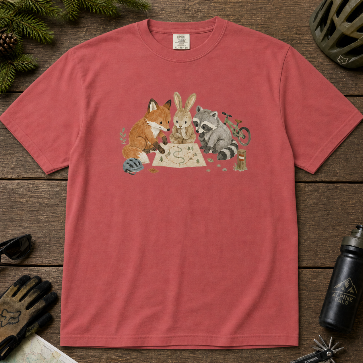 Flat lay of a crimson red t-shirt featuring a cute woodland animal trail-planning graphic with a fox, rabbit, raccoon, map, helmet, bike, and trail marker.