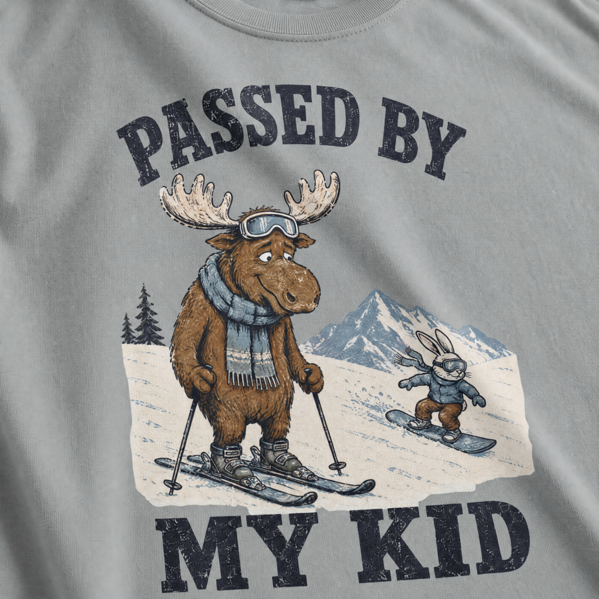 Close-up of a granite gray t-shirt with a vintage “Passed By My Kid” design showing an embarrassed moose skier being passed by a tiny rabbit snowboarder.