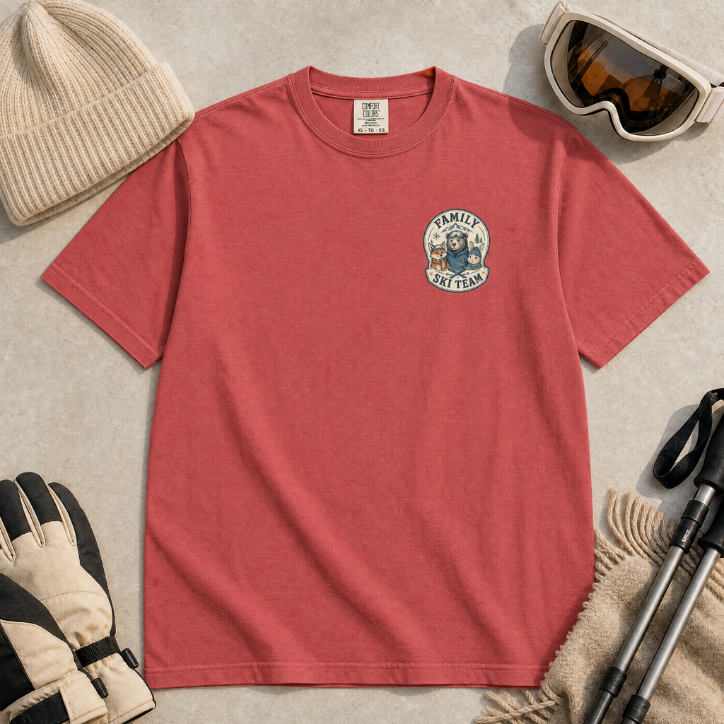 Flat lay of a crimson red t-shirt featuring a small chest “Family Ski Team” badge graphic with three winter animal characters, styled with ski accessories.