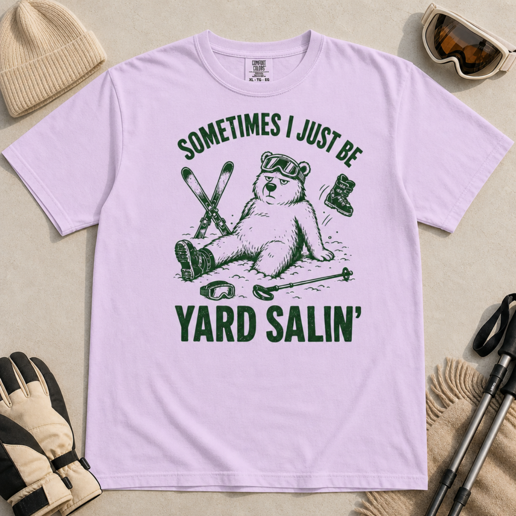 Flat lay of a orchid colored t-shirt featuring a funny “Sometimes I Just Be Yard Salin’” ski design with a tired bear sitting in the snow among scattered gear.