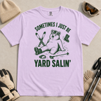Flat lay of a orchid colored t-shirt featuring a funny “Sometimes I Just Be Yard Salin’” ski design with a tired bear sitting in the snow among scattered gear.