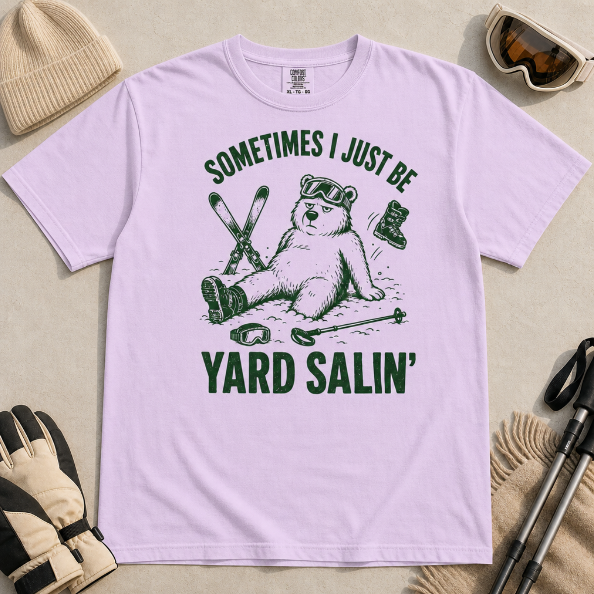 Flat lay of a orchid colored t-shirt featuring a funny “Sometimes I Just Be Yard Salin’” ski design with a tired bear sitting in the snow among scattered gear.