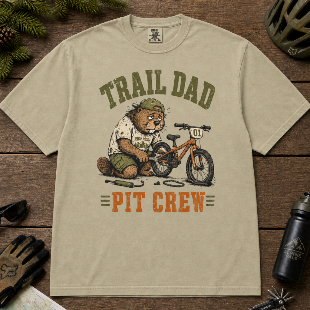 Flat lay of a khaki t-shirt featuring a “Trail Dad Pit Crew” mountain biking design with a tired beaver fixing a kid’s bike, styled with trail accessories.