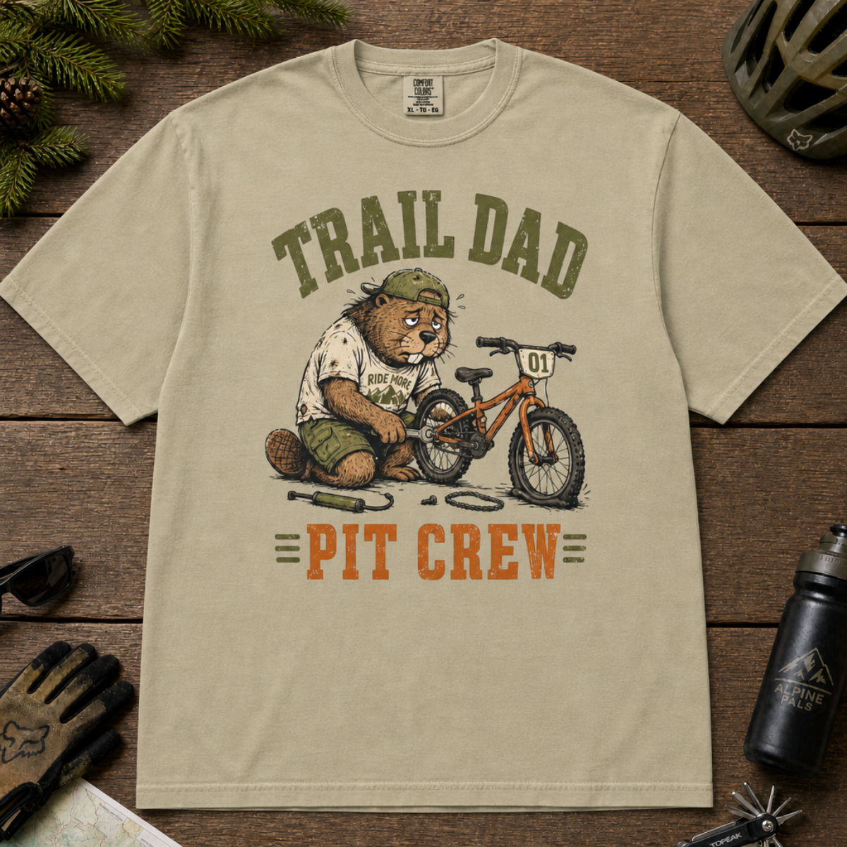 Flat lay of a khaki t-shirt featuring a “Trail Dad Pit Crew” mountain biking design with a tired beaver fixing a kid’s bike, styled with trail accessories.