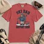 Flat lay of a crimson red t-shirt featuring a tired bear carrying skis and ski gear, with “Ski Dad Support Crew” text.