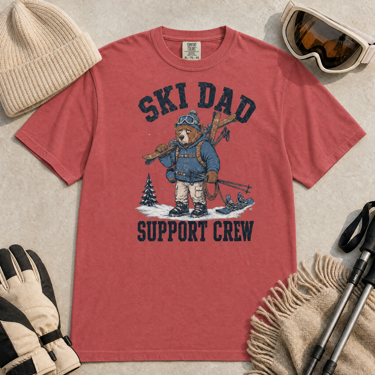 Flat lay of a crimson red t-shirt featuring a tired bear carrying skis and ski gear, with “Ski Dad Support Crew” text.