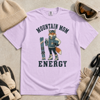 Flat lay of an orchid purple t-shirt featuring a “Mountain Mom Energy” fox ski graphic, styled with winter accessories like goggles, gloves, and poles.