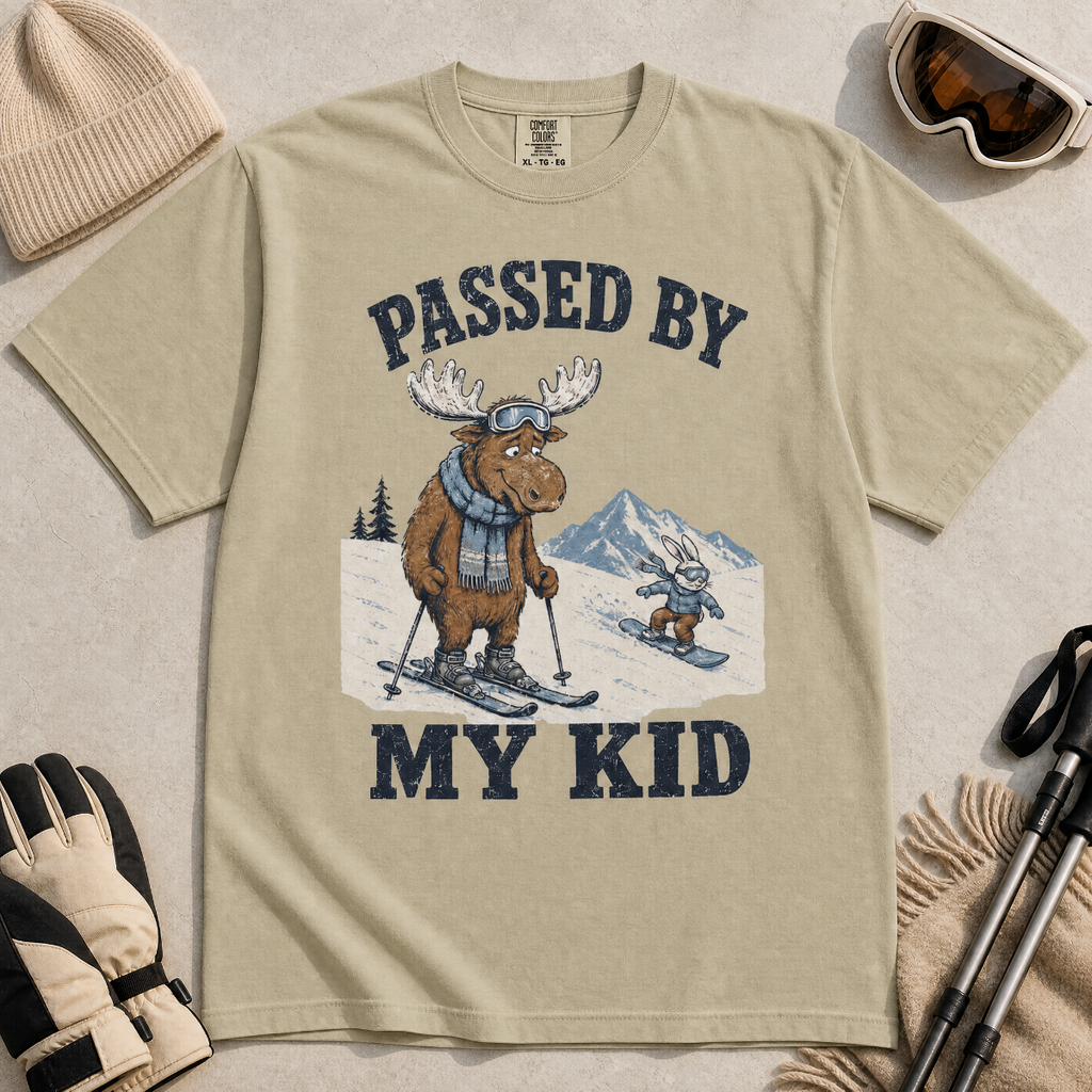 Flat lay of a khaki t-shirt featuring a funny “Passed By My Kid” ski design with a humbled moose watching a small rabbit snowboarder pass by.