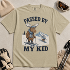 Flat lay of a khaki t-shirt featuring a funny “Passed By My Kid” ski design with a humbled moose watching a small rabbit snowboarder pass by.