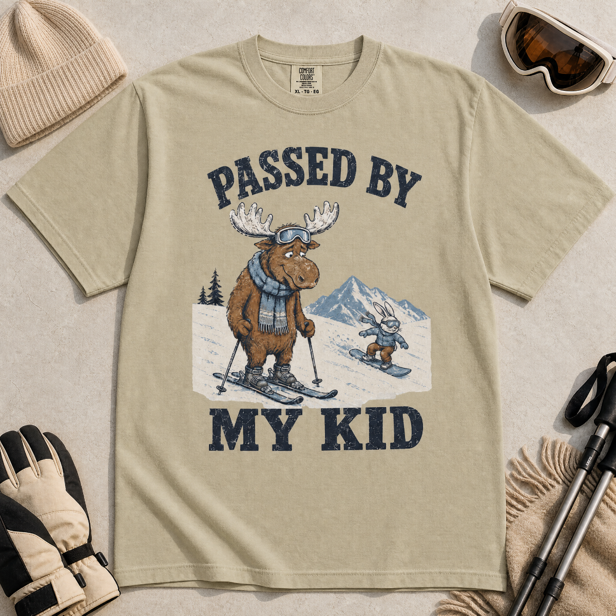 Flat lay of a khaki t-shirt featuring a funny “Passed By My Kid” ski design with a humbled moose watching a small rabbit snowboarder pass by.