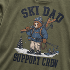 Close-up of a moss green t-shirt with a vintage ski dad design showing an exhausted bear loaded with skis, poles, and snow gear.