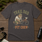 Flat lay of a graphite gray t-shirt featuring a “Trail Dad Pit Crew” mountain biking design with a tired beaver fixing a kid’s bike, styled with trail accessories.