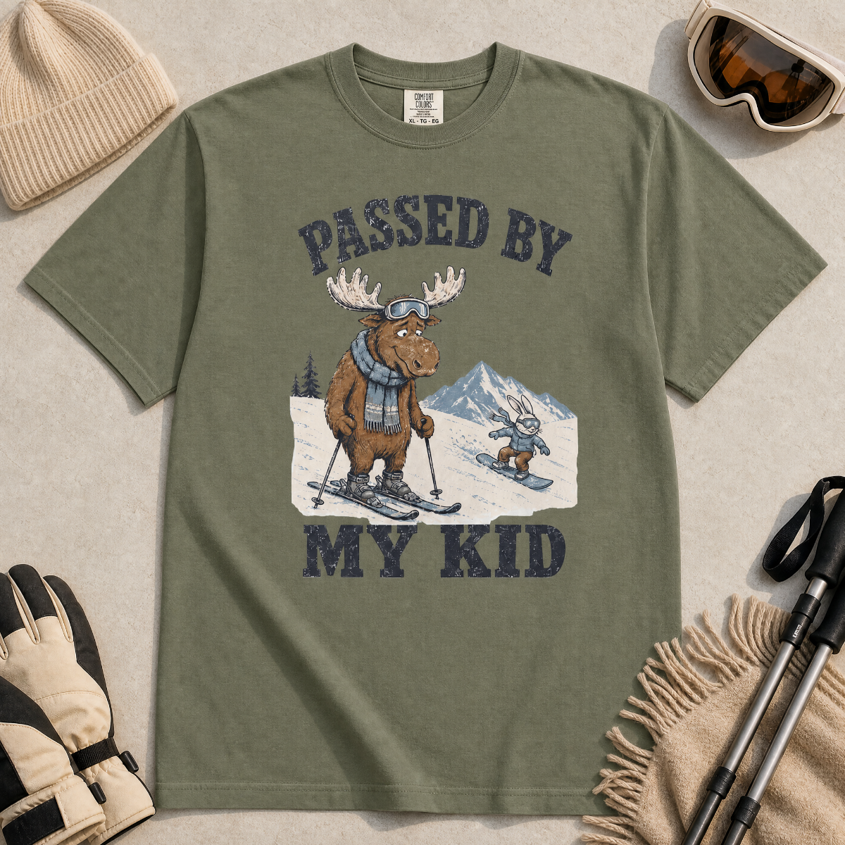 Flat lay of a moss green t-shirt featuring a funny “Passed By My Kid” ski design with a humbled moose watching a small rabbit snowboarder pass by.
