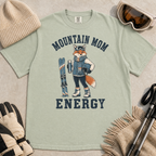 Flat lay of a bay green t-shirt featuring a “Mountain Mom Energy” fox ski graphic, styled with winter accessories like goggles, gloves, and poles.