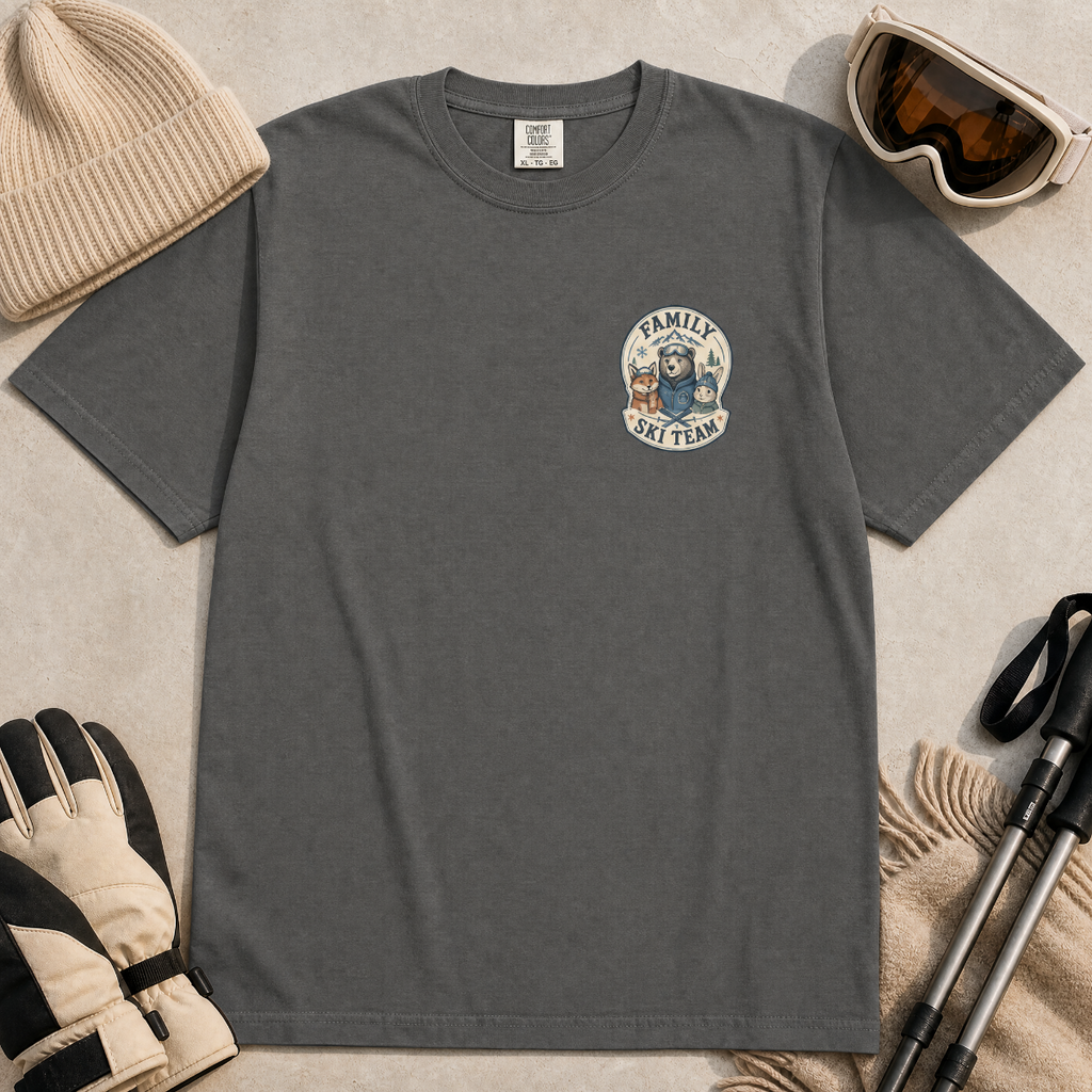 Flat lay of a pepper gray t-shirt featuring a small chest “Family Ski Team” badge graphic with three winter animal characters, styled with ski accessories.