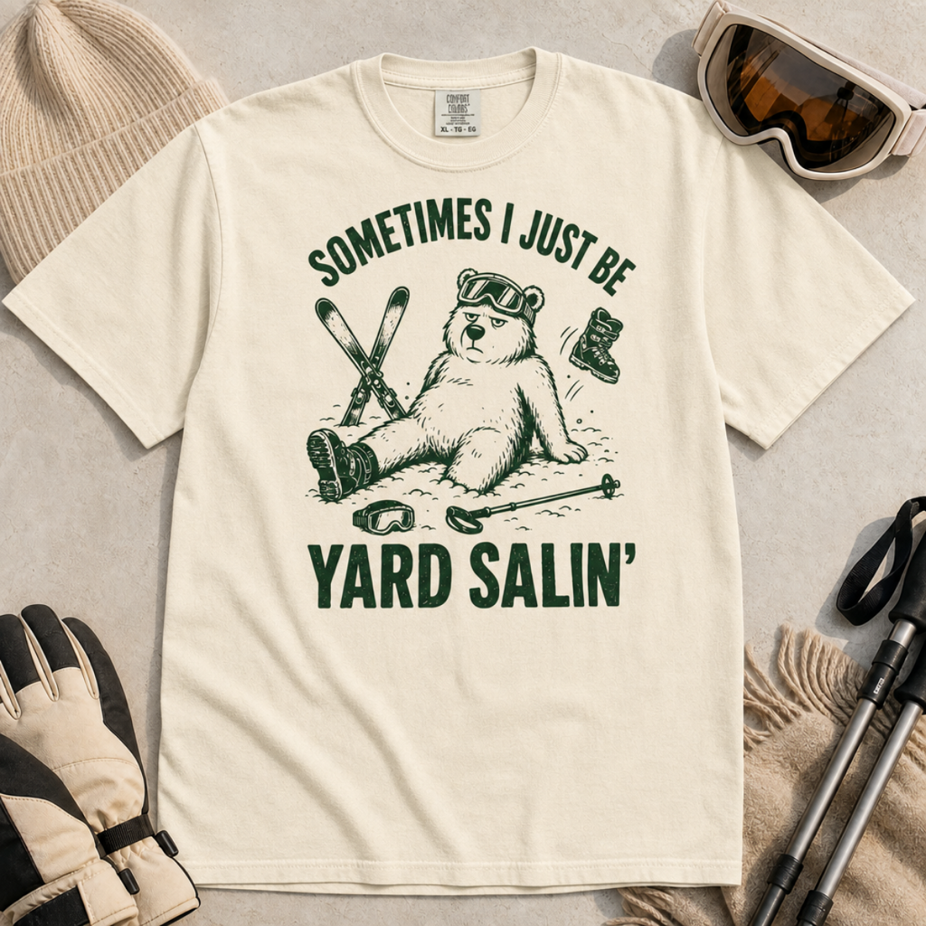 Flat lay of an ivory white t-shirt featuring a funny “Sometimes I Just Be Yard Salin’” ski design with a tired bear sitting in the snow among scattered gear.