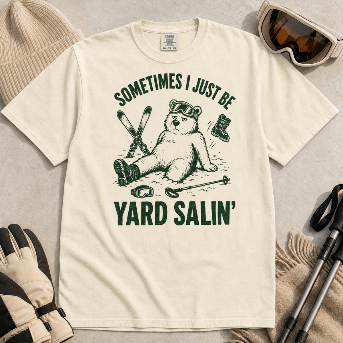 Flat lay of an ivory white t-shirt featuring a funny “Sometimes I Just Be Yard Salin’” ski design with a tired bear sitting in the snow among scattered gear.