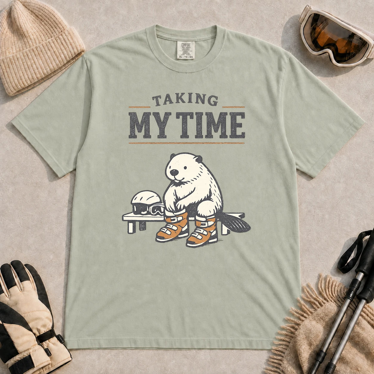 Bay tee featuring a relaxed beaver sitting on a bench in ski boots beside a helmet and goggles, with the phrase “Taking My Time.”