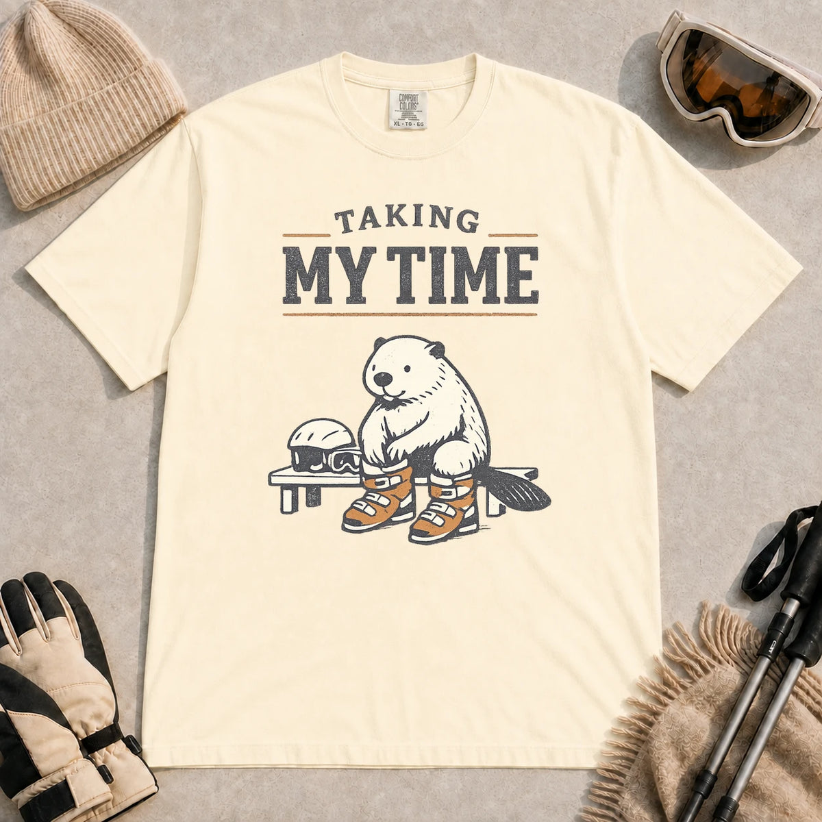 Ivory tee featuring a relaxed beaver sitting on a bench in ski boots beside a helmet and goggles, with the phrase “Taking My Time.”